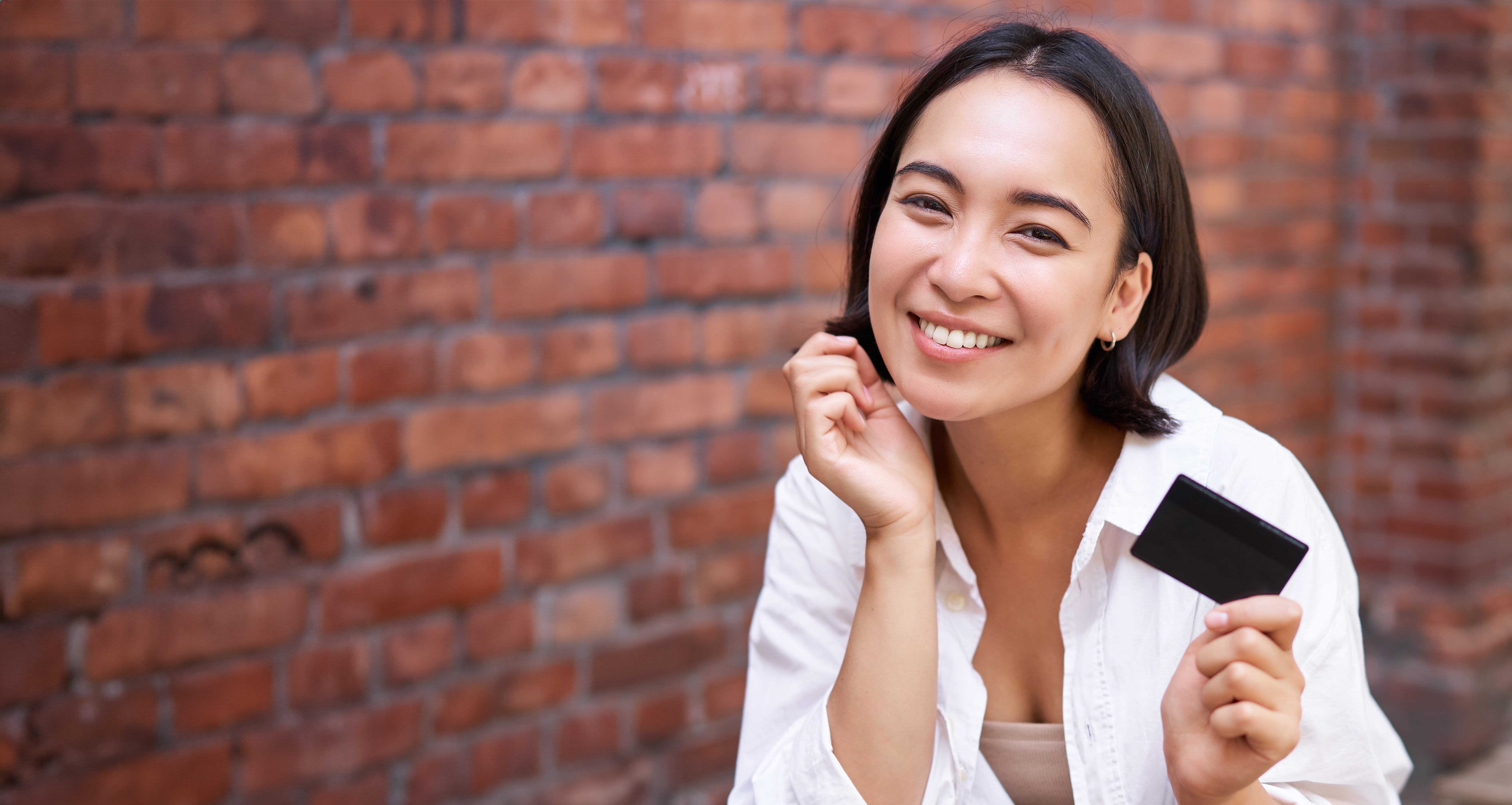 Smiling woman holding a credit card in front of a brick wall.