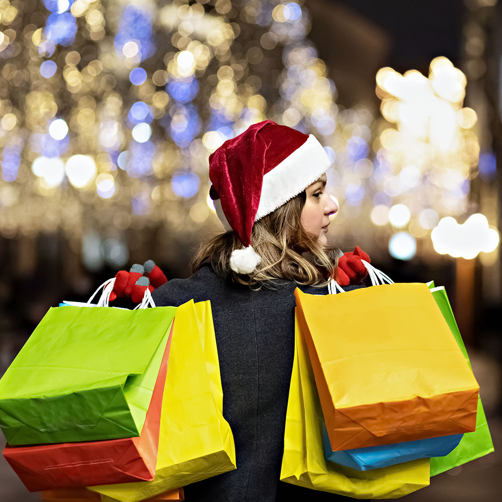 A woman with long hair and a santa hat near the window of a city store with purchases paper bags