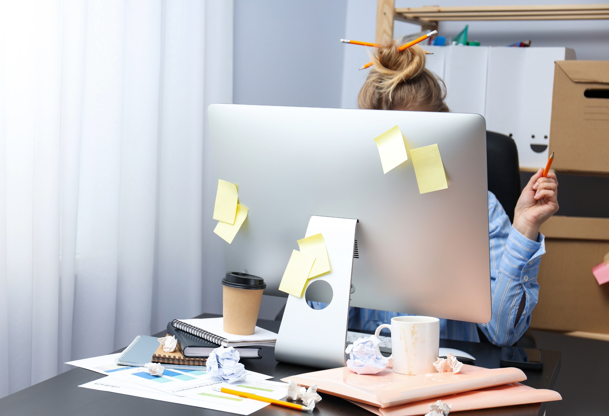 photo of woman behind computer monitor with sticky notes, coffee and papers everywhere