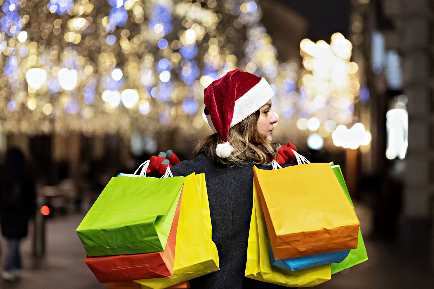 A woman with long hair and a santa hat near the window of a city store with purchases paper bags