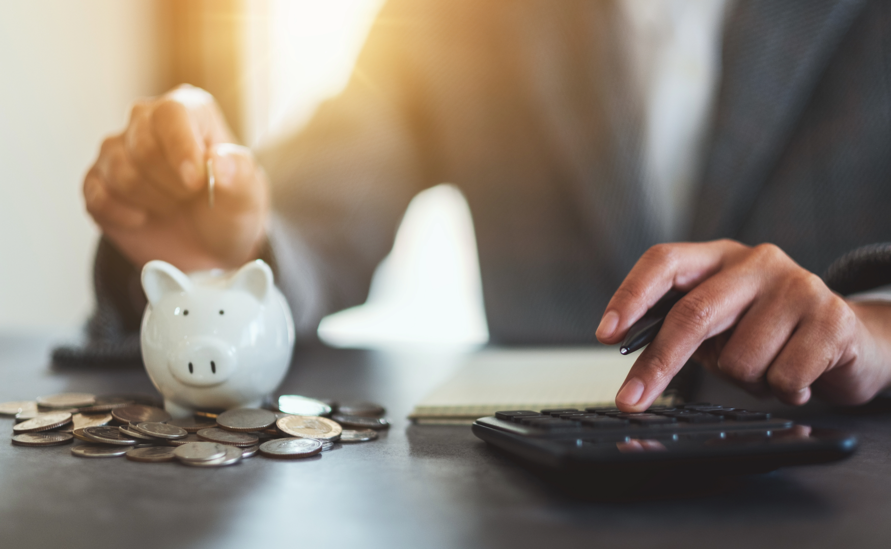 close up of man at desk with calculator under one hand and a piggy bank with coins under another