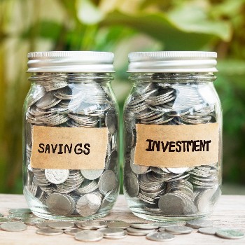 Two glass jars filled with coins, sitting on a table, one labeled savings and one labeled investment