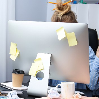 photo of woman behind computer monitor with sticky notes, coffee and papers everywhere