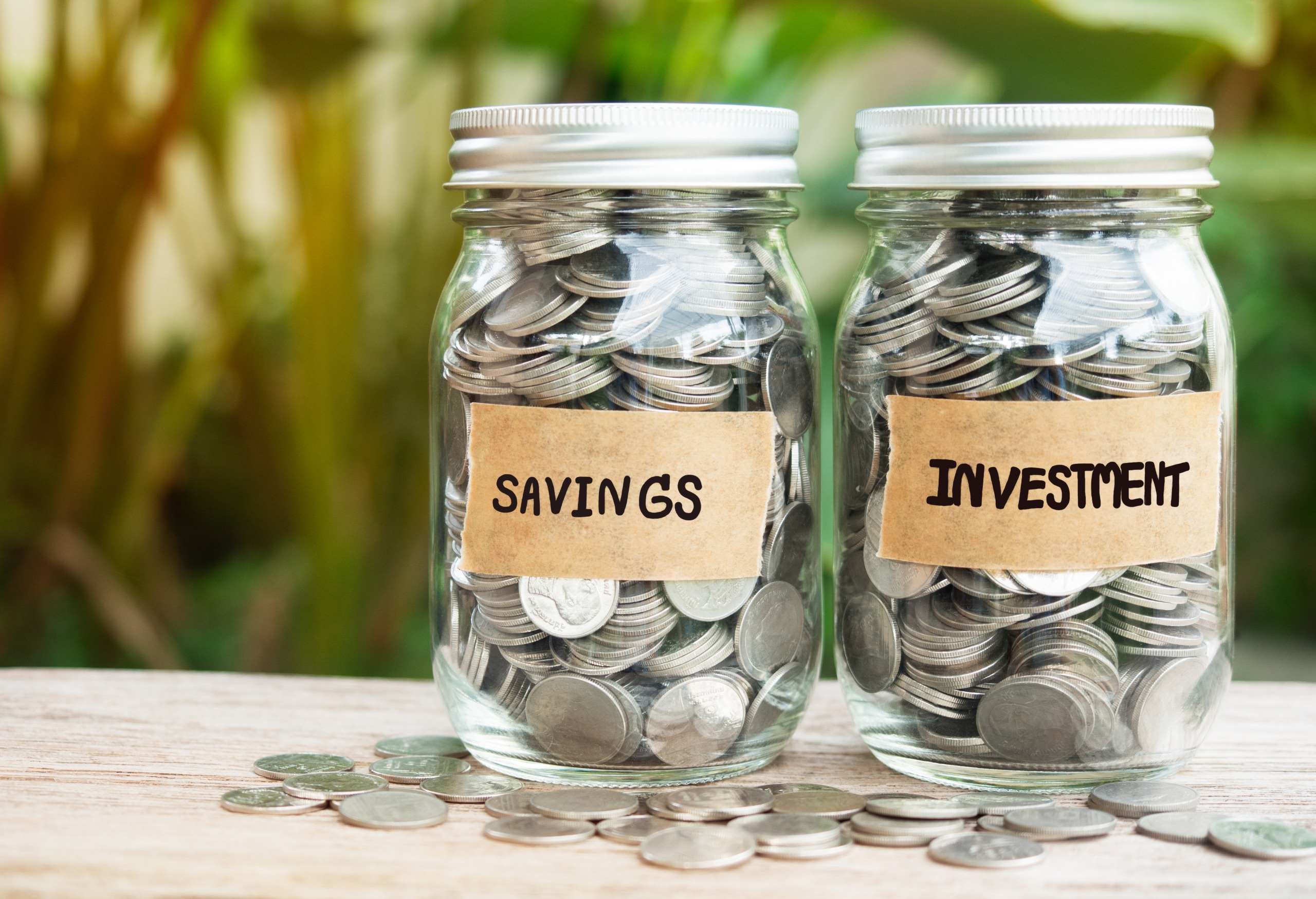 Two glass jars filled with coins, sitting on a table, one labeled savings and one labeled investment