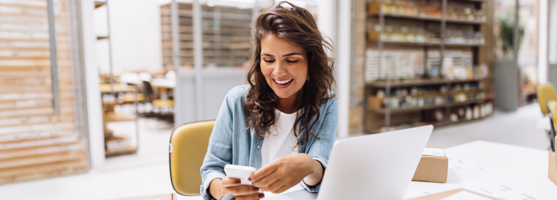 woman looking at a mobile phone while working on a laptop