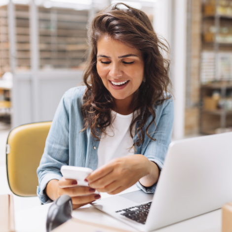 woman looking at a mobile phone while working on a laptop